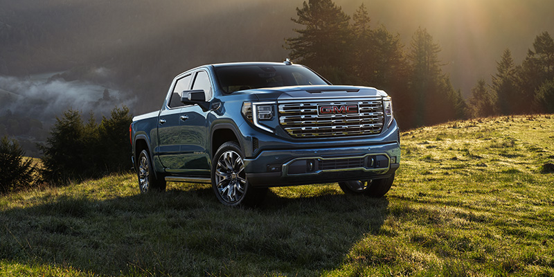 A blue-grey 2026 GMC Sierra 1500 is shown off with the glancing sunshine as it drives over a green mountain hillside meadow.