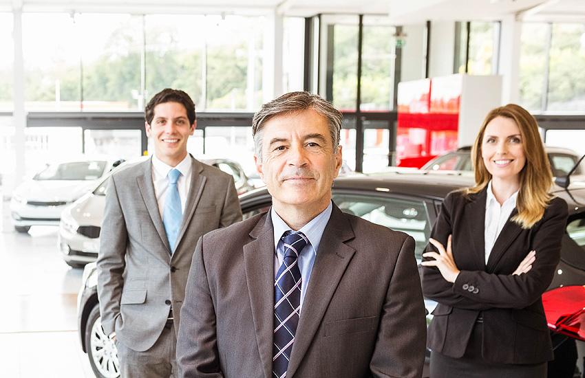 3 smiling salespersons stand in sunlilt GMC showroom, ready to serve new vehicle shoppers.